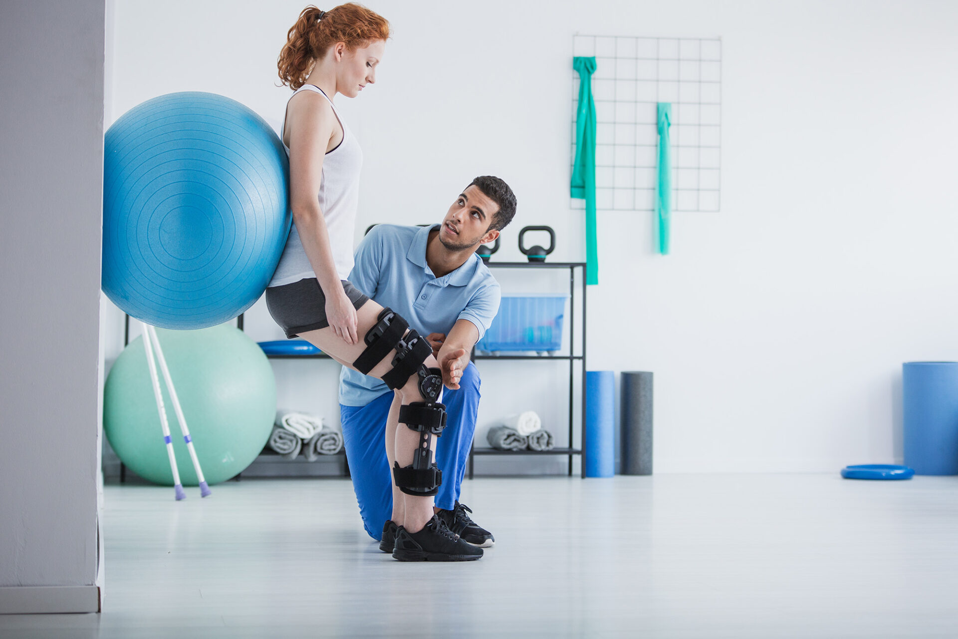 Schneller zurück im Alltag mit der Krankentagegeldversicherung Junge Frau mit Knieorthesen macht unter Anleitung eines Physiotherapeuten eine Übung mit einem Gymnastikball an der Wand. Symbol für Rehabilitation, Krankentagegeld und gezielte Unterstützung bei Genesung und Wiedereingliederung.