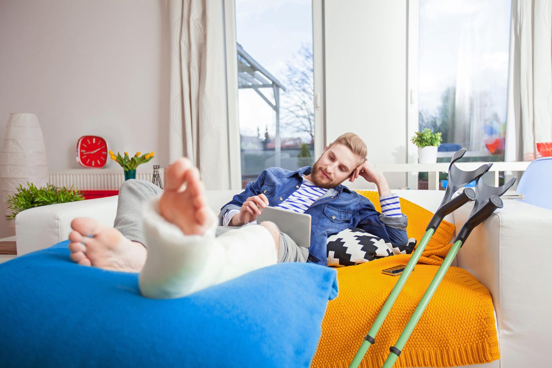 Mann mit Gips am Fuß liegt auf dem Sofa in einem Haus und schau auf ein Tablet. Neben ihm stehen Krücken. Er sieht zufrieden aus.