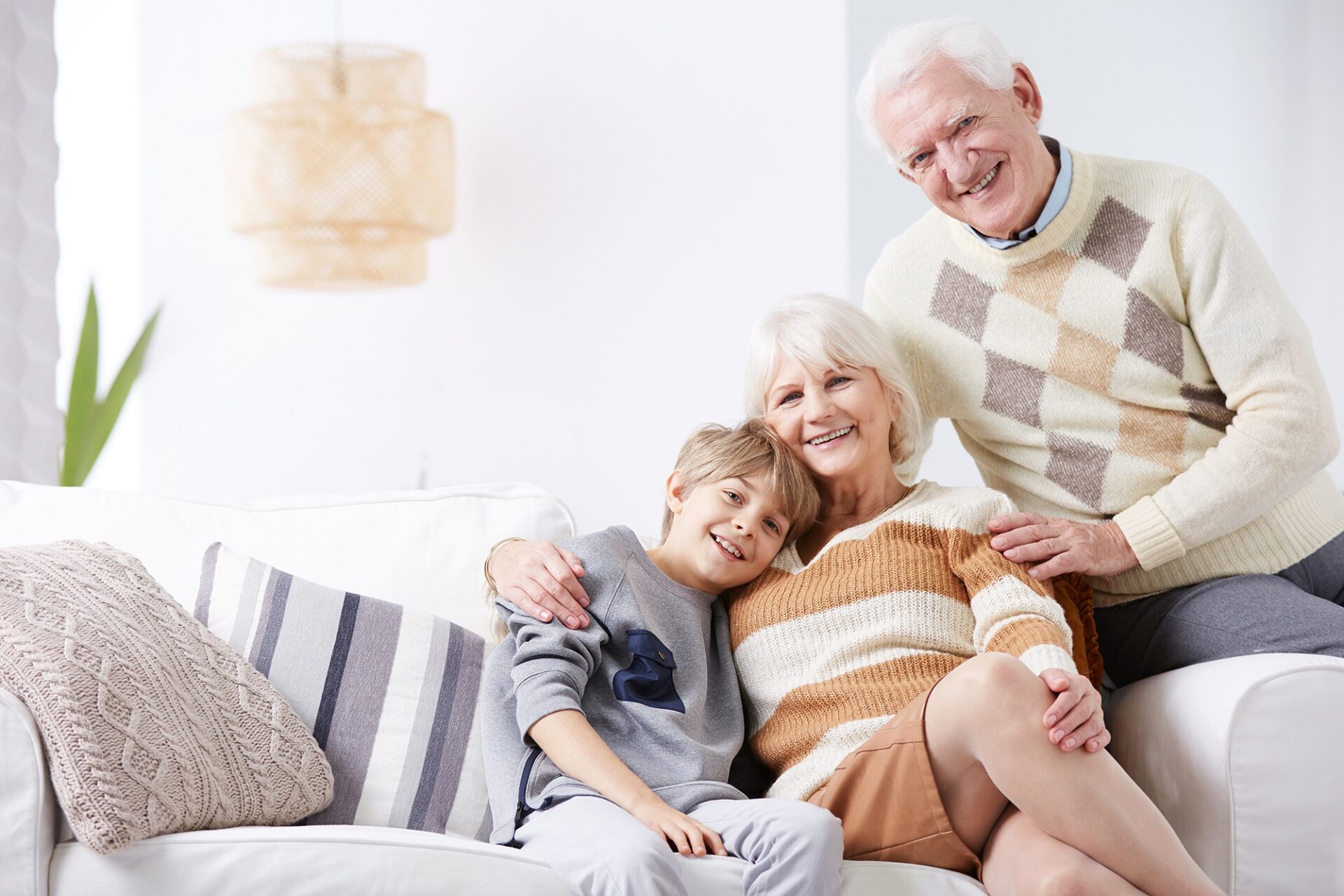 Glückliche Großeltern sitzen mit ihrem Enkelkind auf einem weißen Sofa, alle drei lächeln in die Kamera. Symbol für Geborgenheit, familiäre Nähe und Absicherung im Alter.