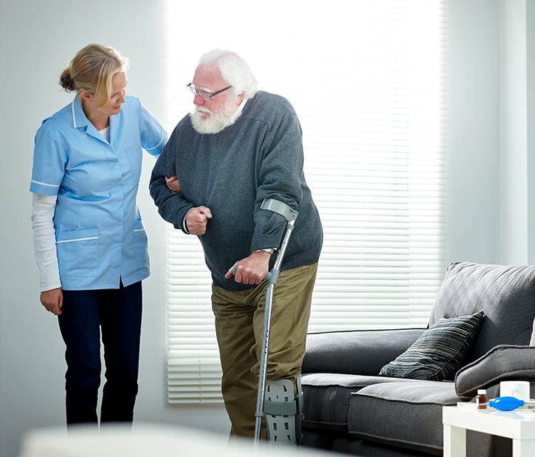 Älterer Mann mit Krücken und Fußschiene wird von einer Pflegerin beim Aufstehen im Wohnzimmer unterstützt. Darstellung von Pflegebedürftigkeit und professioneller Hilfe im Alltag.