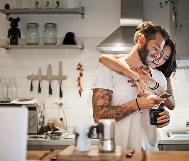 Schneller zurück im Alltag mit der Krankentagegeldversicherung Ein Paar steht in der Küche. Der Mann hält eine Tasse in der Hand und schaut auf sein Handy. Die Frau umarmt ihn von hinten und schaut mit auf das Handy.
